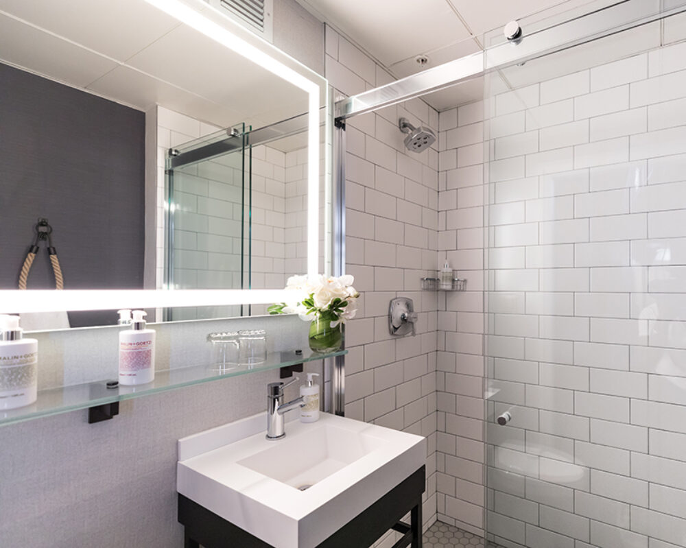 Modern bathroom at Harbor Court Hotel, equipped with a glass-enclosed shower, illuminated vanity mirror, sleek white tiles, and fresh floral decor