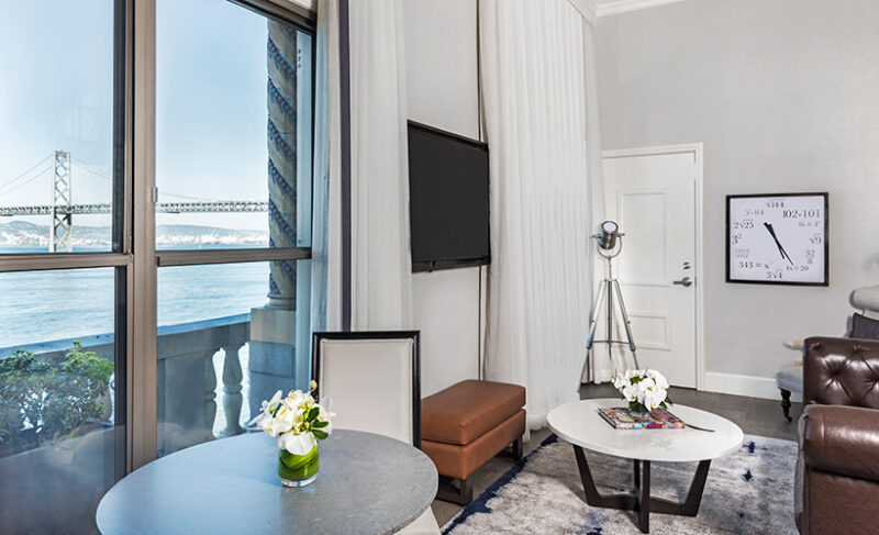 living room of the Skyline Waterfront Penthouse Suite at Harbor Court Hotel, featuring a large window with a stunning view of the San Francisco Bay Bridge, modern furnishings, and elegant decor